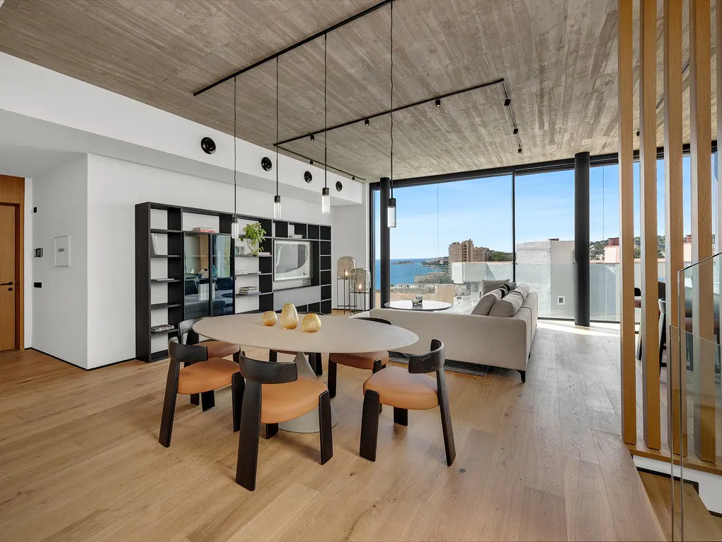 Modern living room with an oval table, chairs, and a large window overlooking the ocean. The room has a concrete ceiling and wood floors.