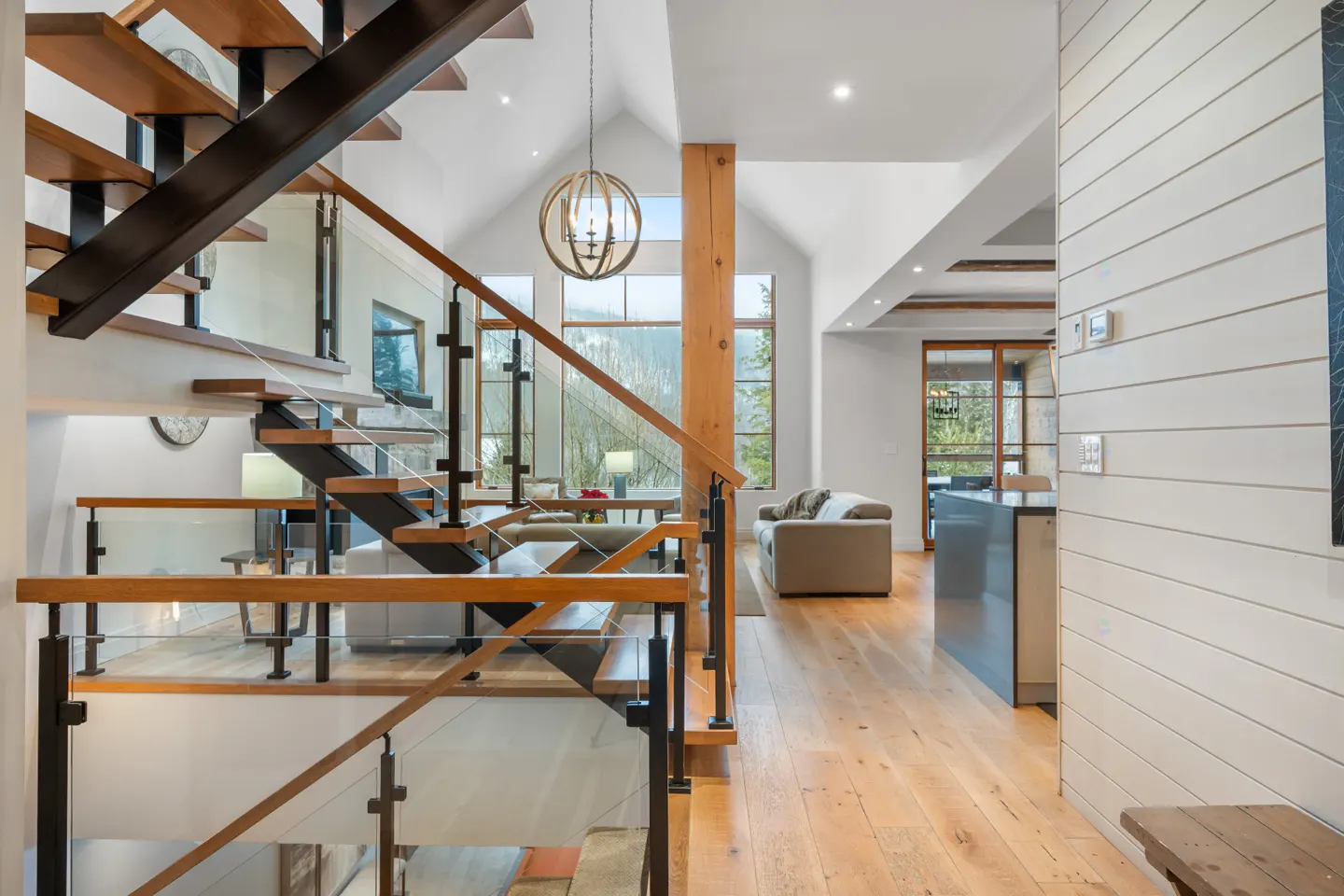 Interior view of a modern home with wood floors, a staircase with glass railings, and a living room with large windows.