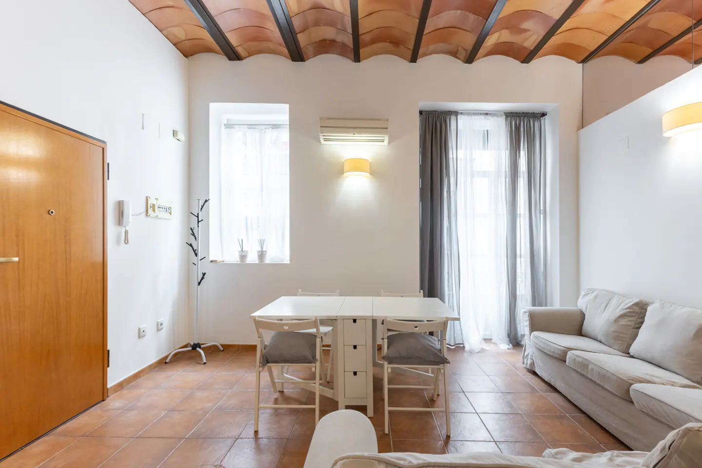 Bright living room with white walls, terracotta tile floor, and a unique terracotta tile ceiling. A white table with chairs sits near a window with gray curtains. A beige sofa is on the right.