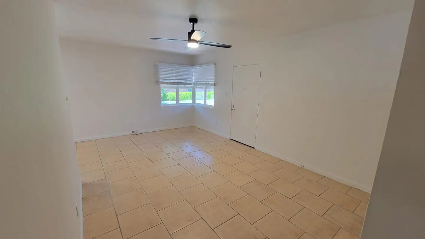 A bright, empty room with beige tile flooring, white walls, a window with blinds, and a ceiling fan.