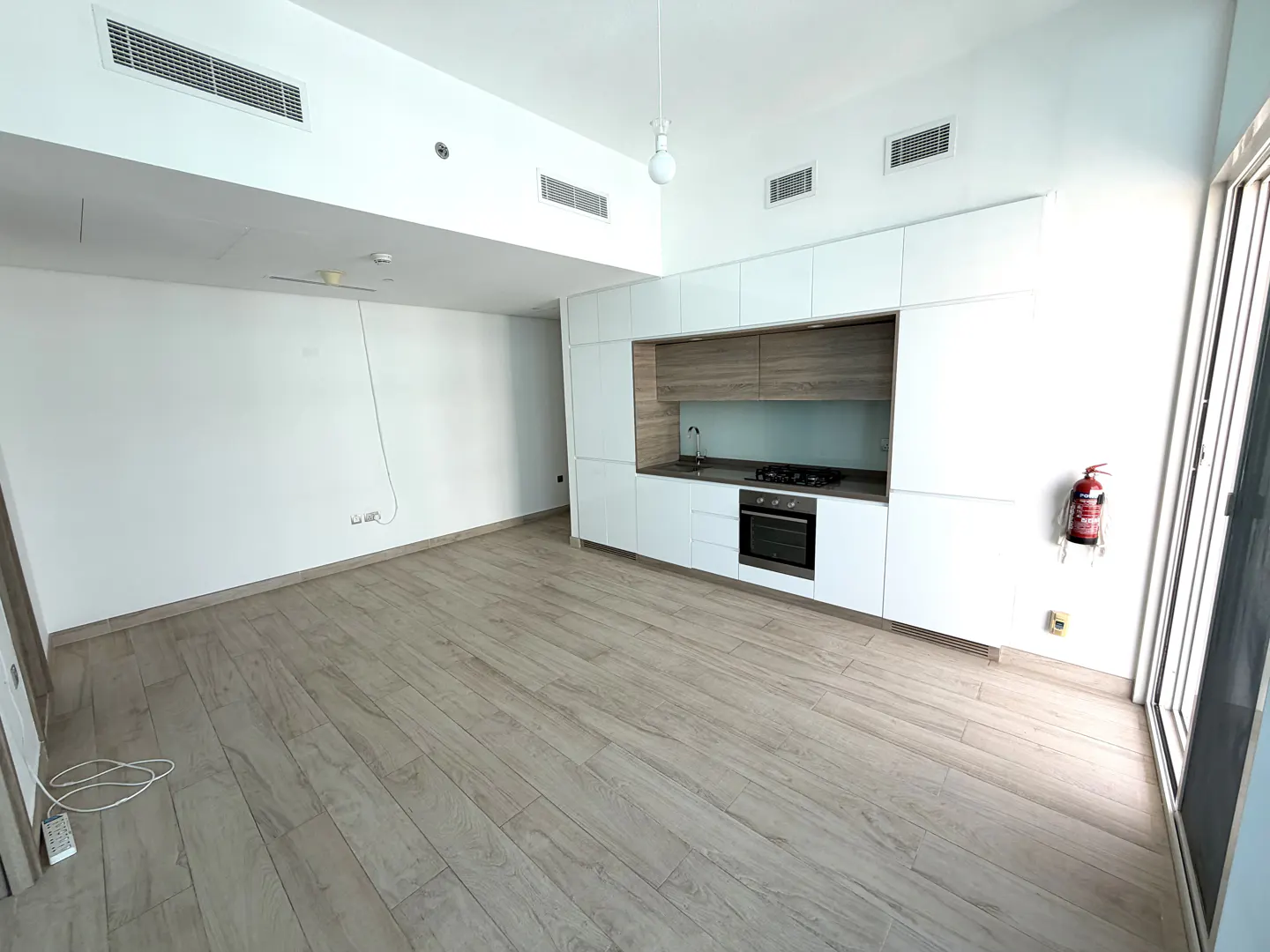 A bright, empty apartment with light wood floors and white walls. A modern kitchen with white cabinets and stainless steel appliances is visible.