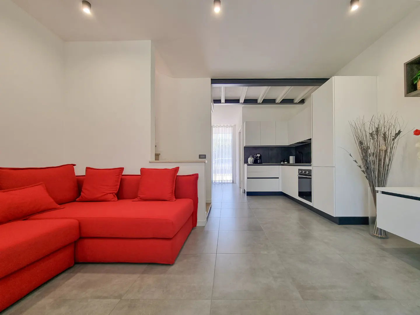 Open-concept living space with a red sectional sofa and a modern white kitchen. Gray tile flooring throughout.