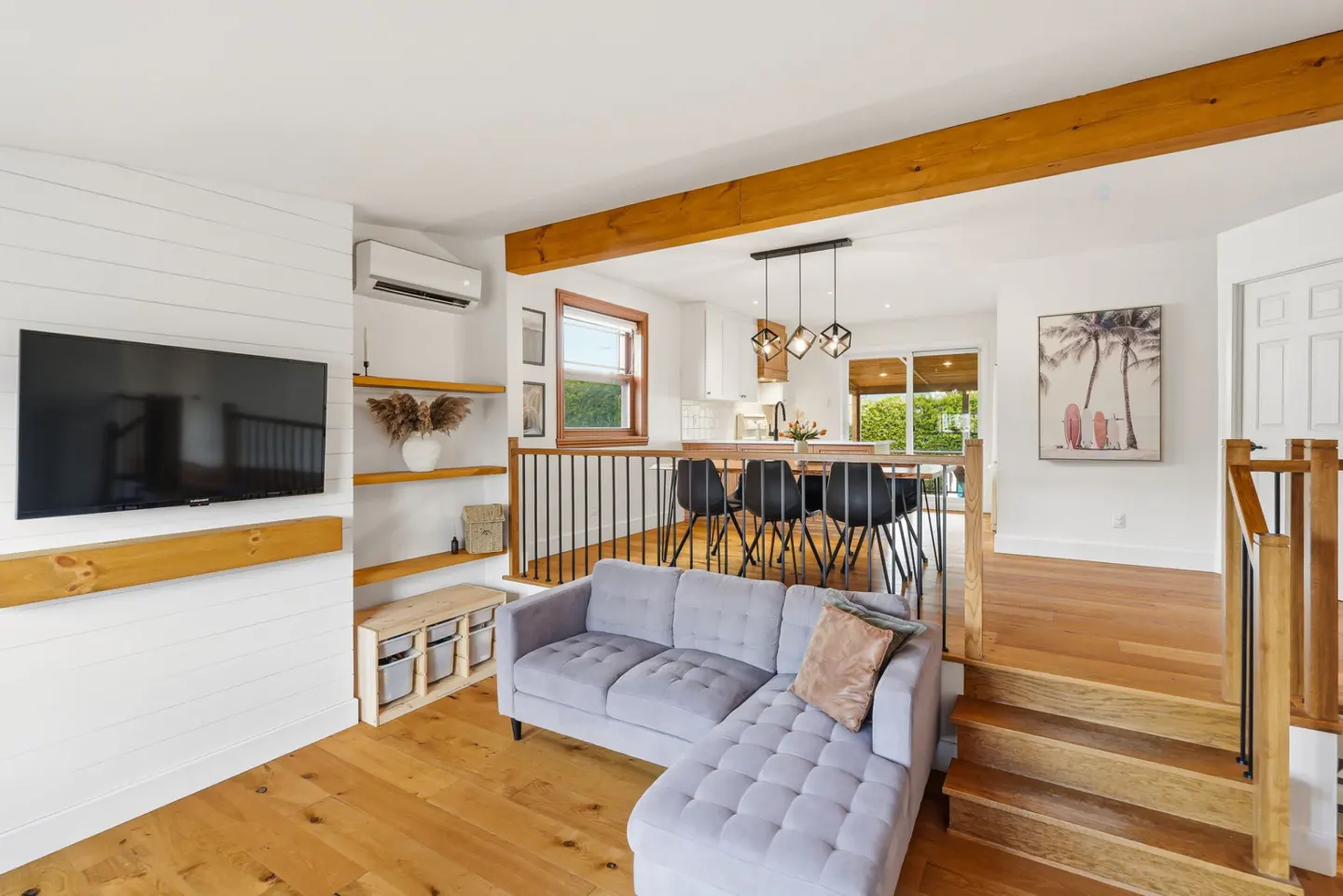 Open-concept living room with a gray sofa, wood floors, and a view of the kitchen. Stairs lead to another level. A TV hangs on the wall.