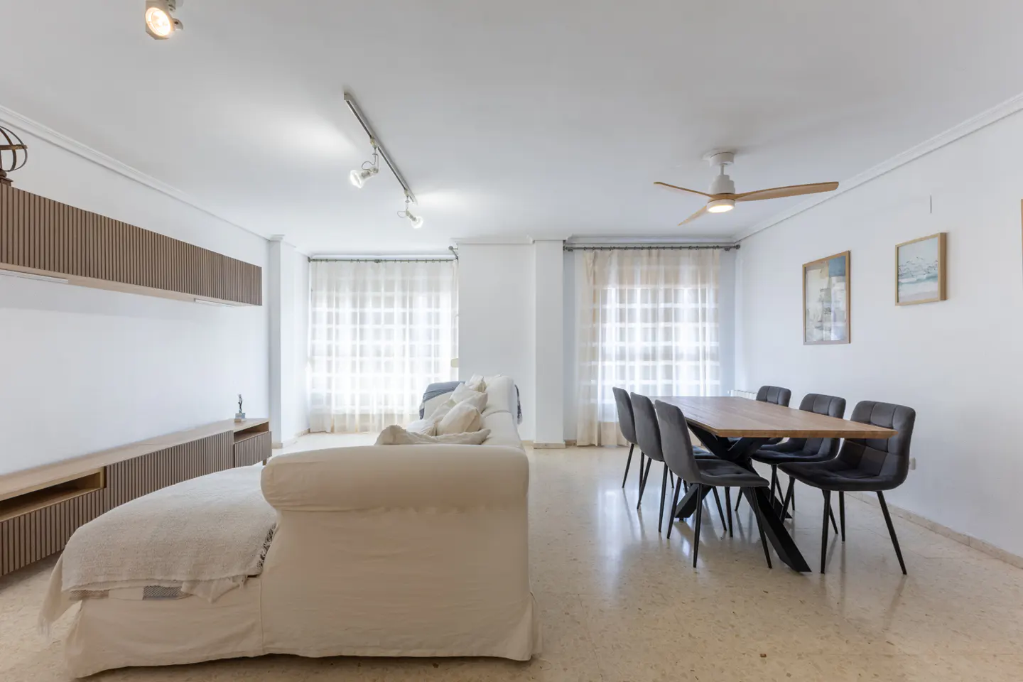 Bright, modern living room with a white sofa, wood dining table with six gray chairs, and built-in wood shelving.