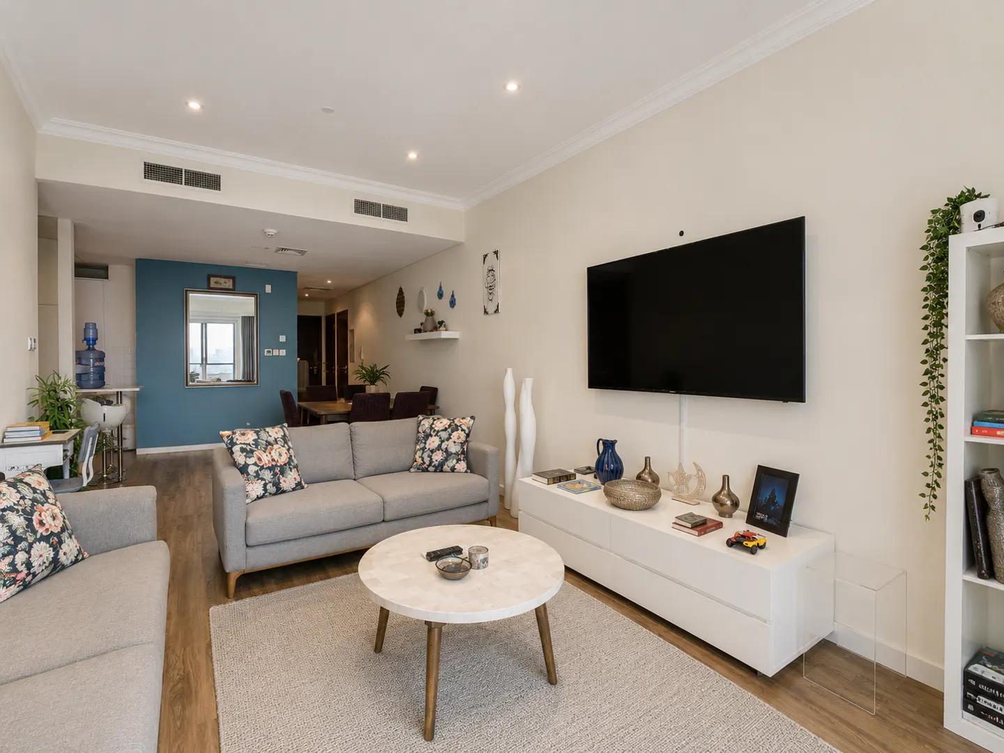 A bright living room with gray sofas, floral pillows, and a round coffee table on a light rug. A large TV hangs above a white console.