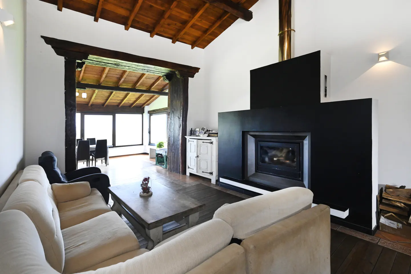 Living room with white sofas, wood ceiling, and black fireplace. Dining room visible through wood-framed opening.