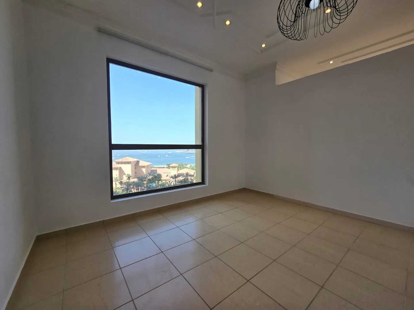 Empty room with beige tile floor, white walls, and a large window showing a coastal view with buildings and the ocean.
