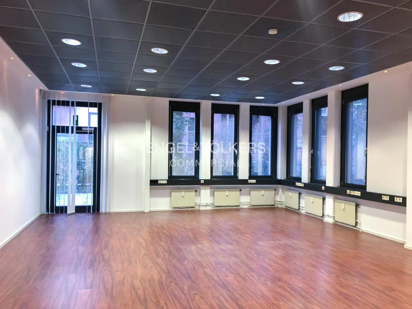 Empty office space with wood floors, white walls, and a gray ceiling with recessed lighting. Windows line one wall, and a glass door is on another.
