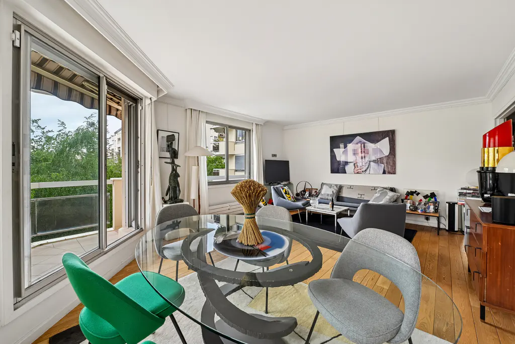 Bright living room with a glass table, green and gray chairs, and a balcony with trees visible through the open doors.