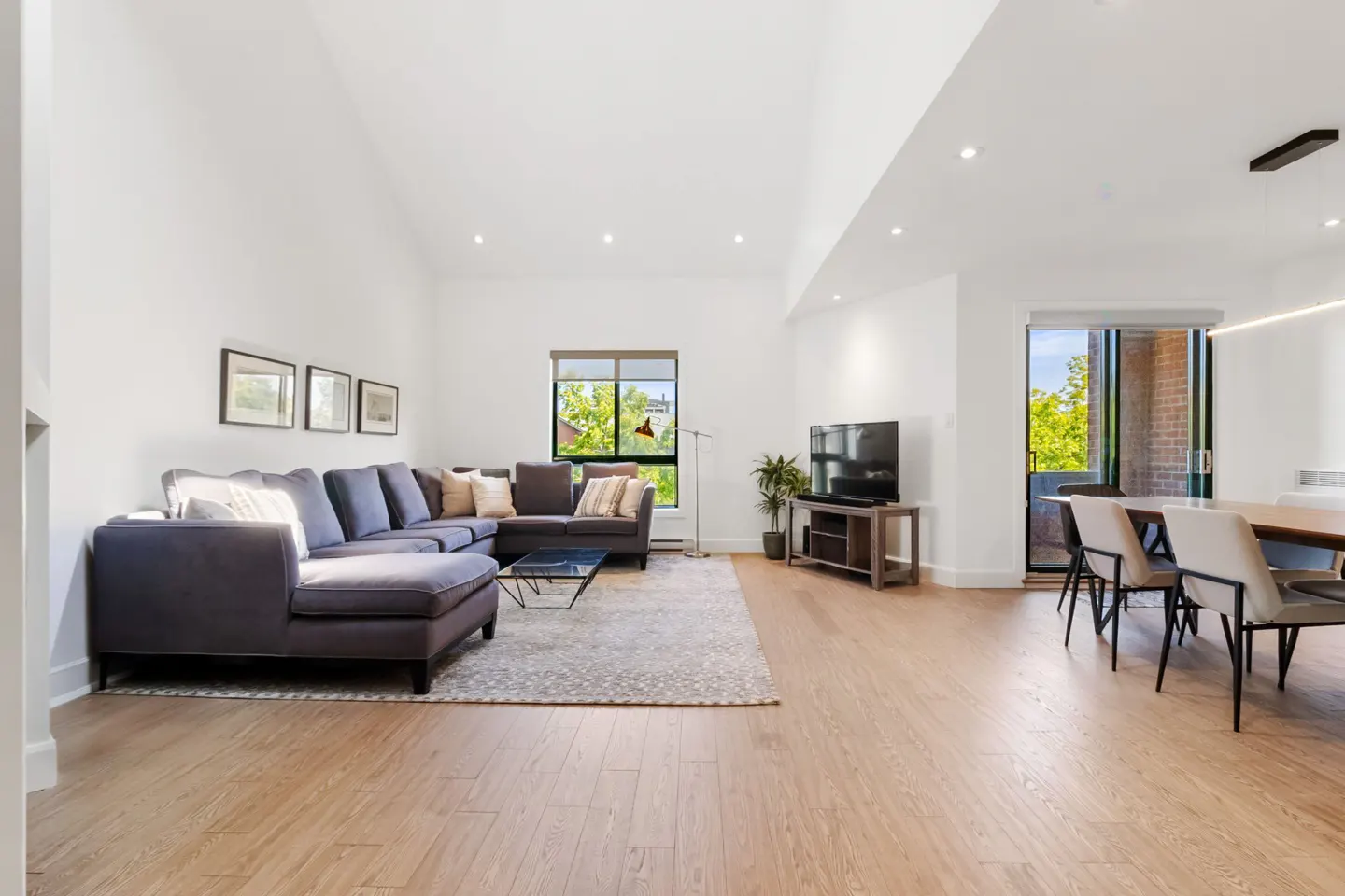Bright, open-concept living room with hardwood floors, a gray sectional sofa, and a dining area with a table and chairs.