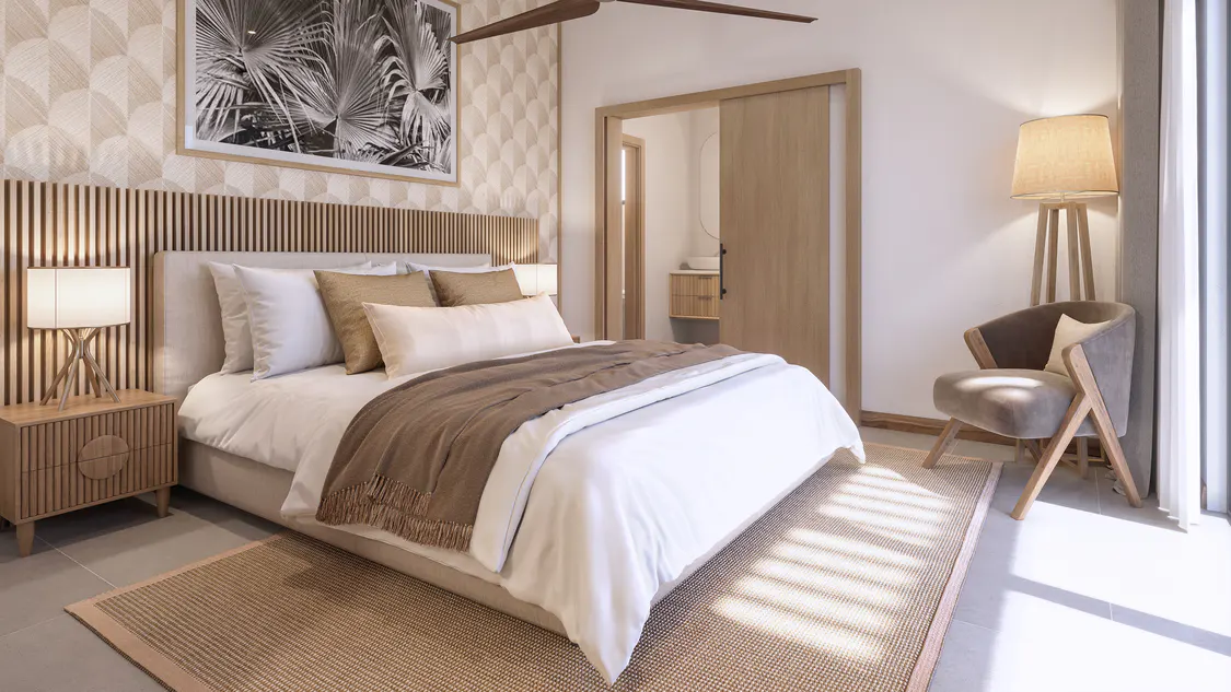 A bright bedroom with a bed, nightstand, lamp, chair, and a sliding door to a bathroom. The bed has white linens and a brown throw.