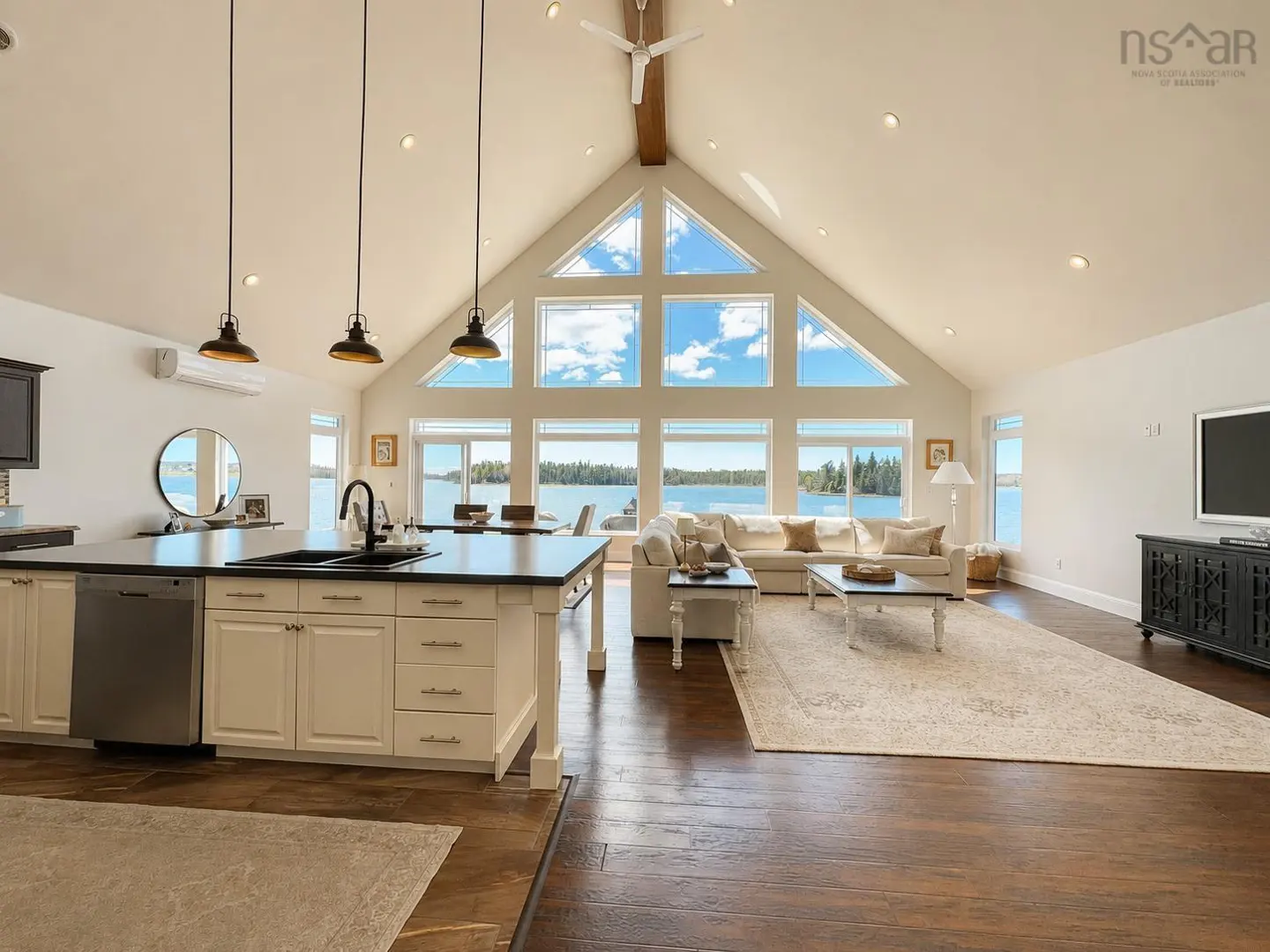 Open-concept living area with white kitchen island, vaulted ceilings, and large windows overlooking a scenic lake.
