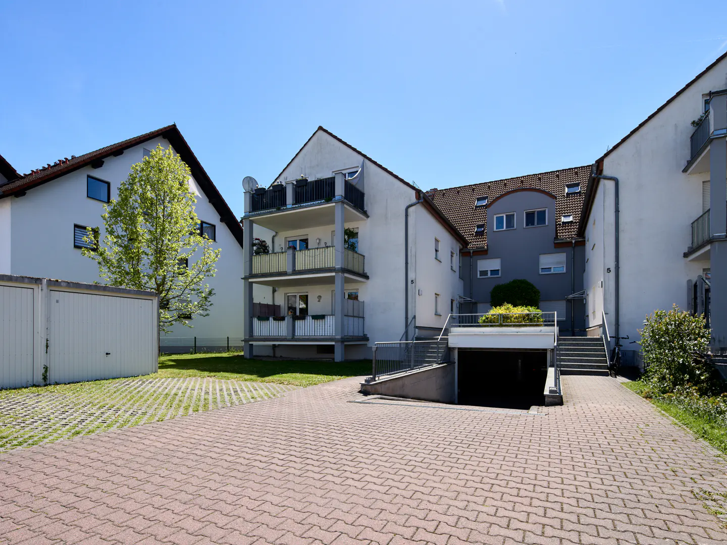 Exterior view of a three-story apartment building with balconies, a garage, and a brick driveway on a sunny day.