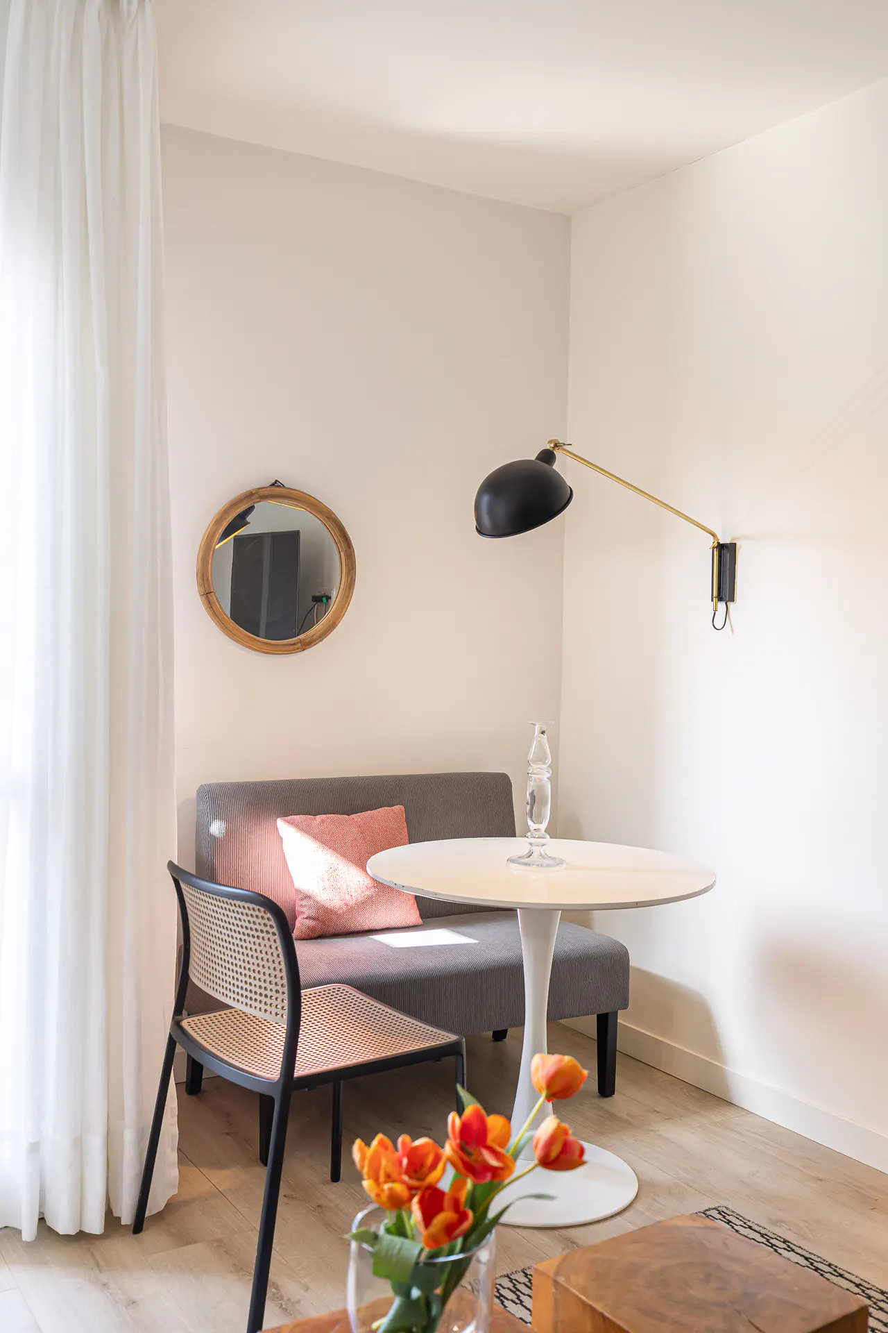 A bright living room with a gray sofa, pink pillow, round table, black lamp, and orange tulips.