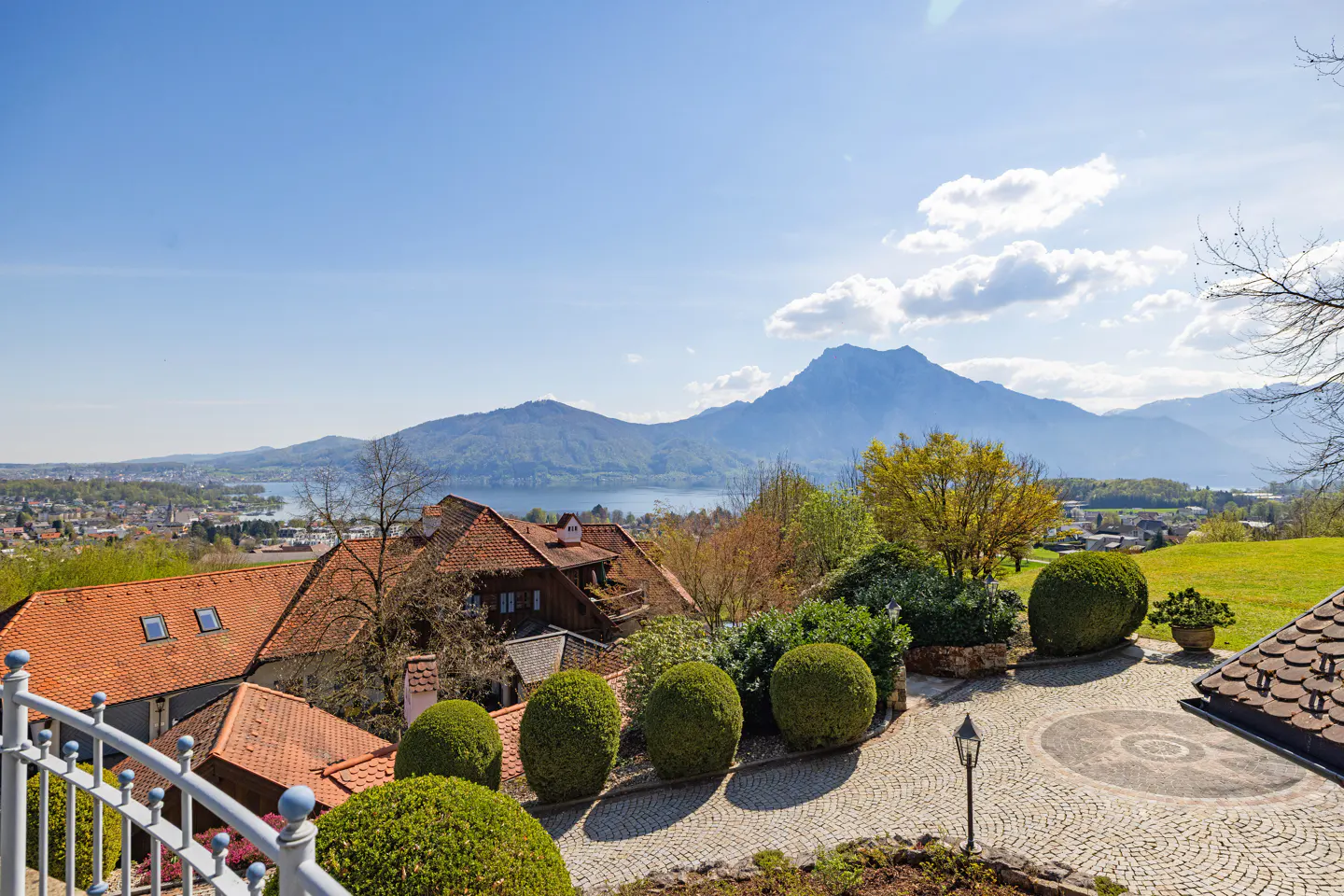 Scenic view of rooftops with orange tiles, green bushes, and a stone driveway. Mountains and a lake are visible in the background under a blue sky.