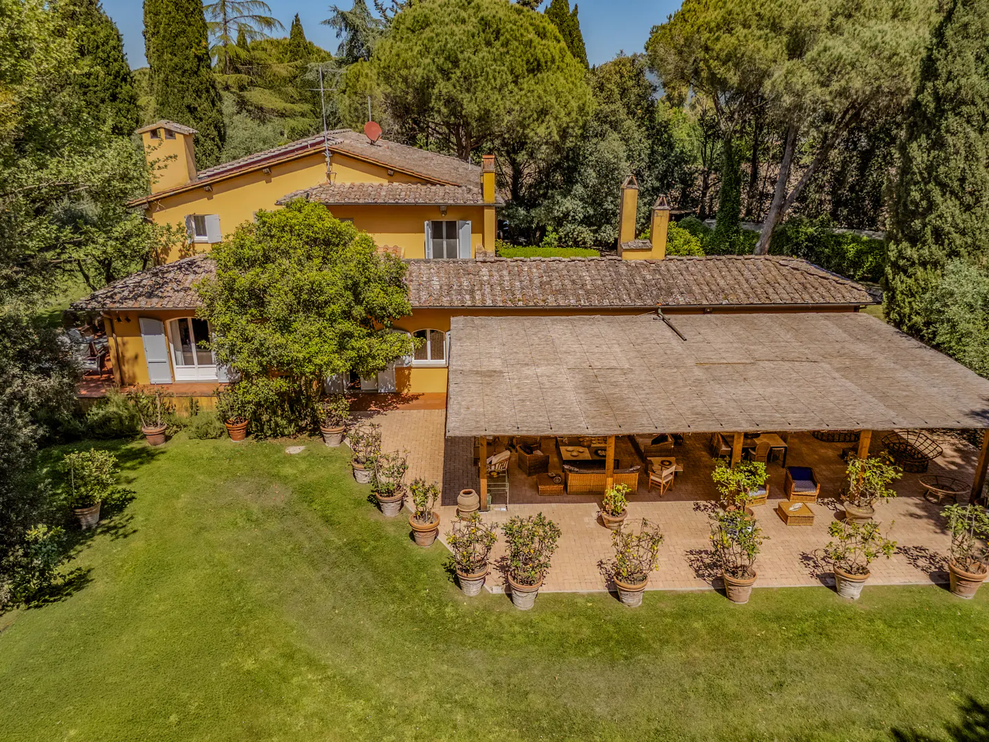 Aerial view of a yellow Tuscan-style villa with a covered patio, surrounded by green trees and a lawn.