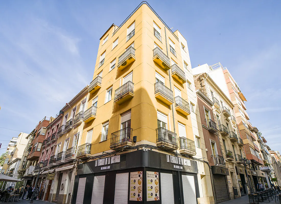 Street view of a tall yellow apartment building with black balconies and a restaurant on the ground floor.