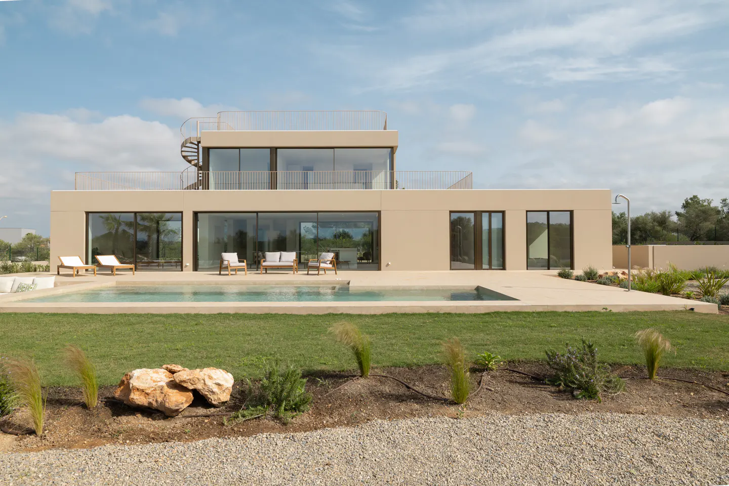 Modern beige two-story house with a pool, lawn, and gravel driveway under a blue sky. Spiral staircase leads to the rooftop.