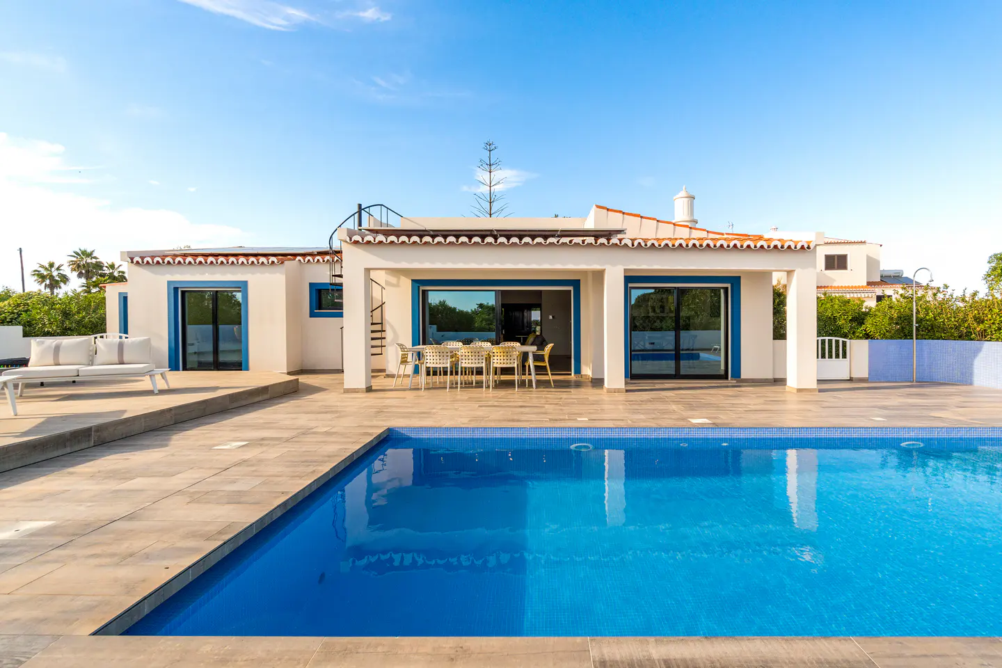 Exterior view of a white house with blue trim, a pool, and outdoor furniture under a sunny blue sky.