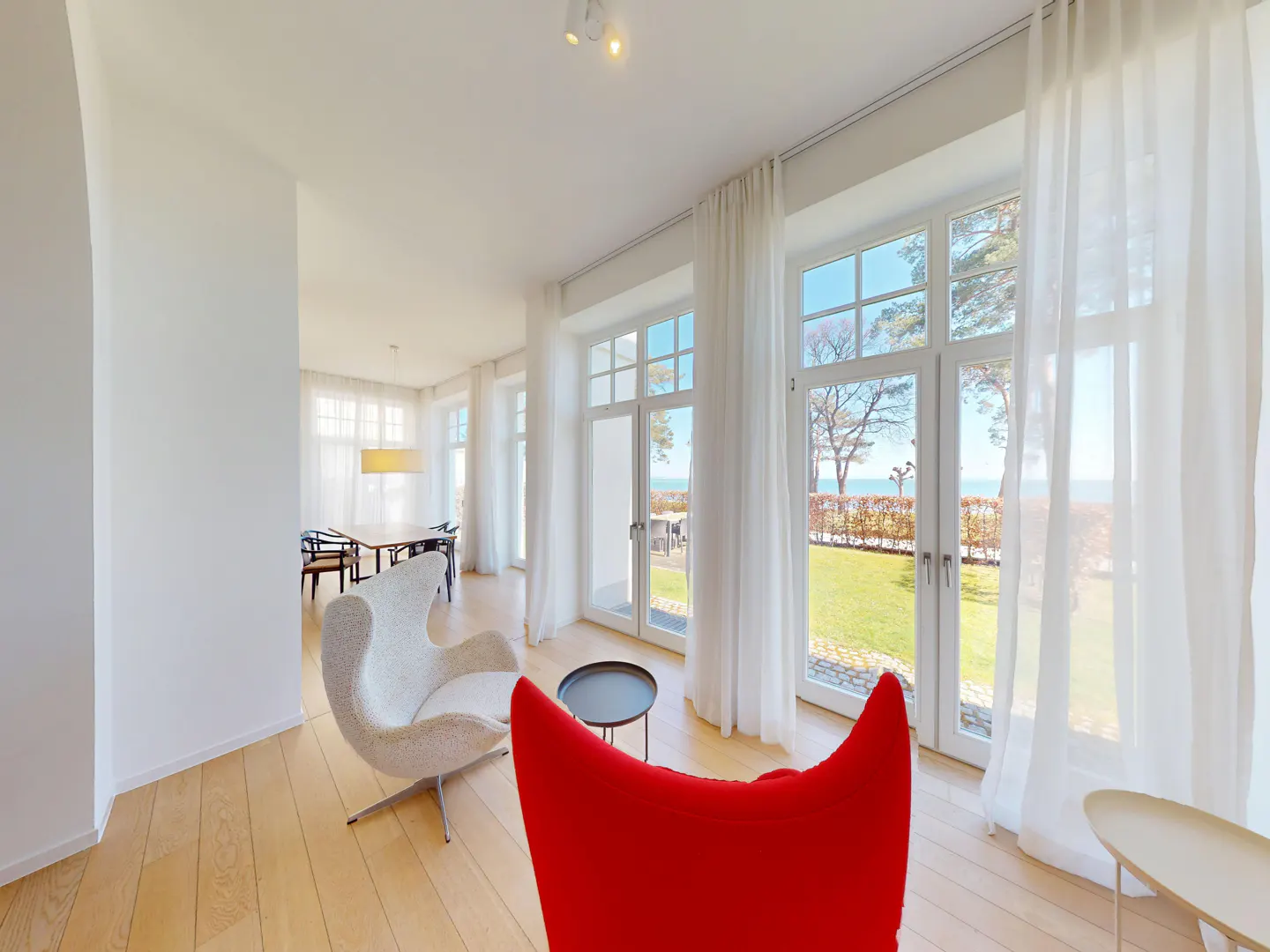 Bright living room with wood floors, white walls, and large windows with sheer white curtains. Red and white chairs face the ocean view.