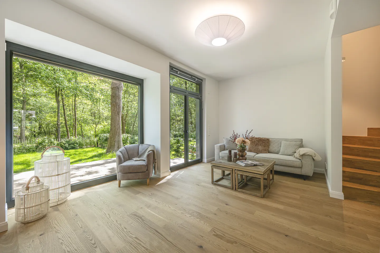 Bright living room with wood floors, large windows overlooking green trees, gray sofa, chair, and wooden coffee table.