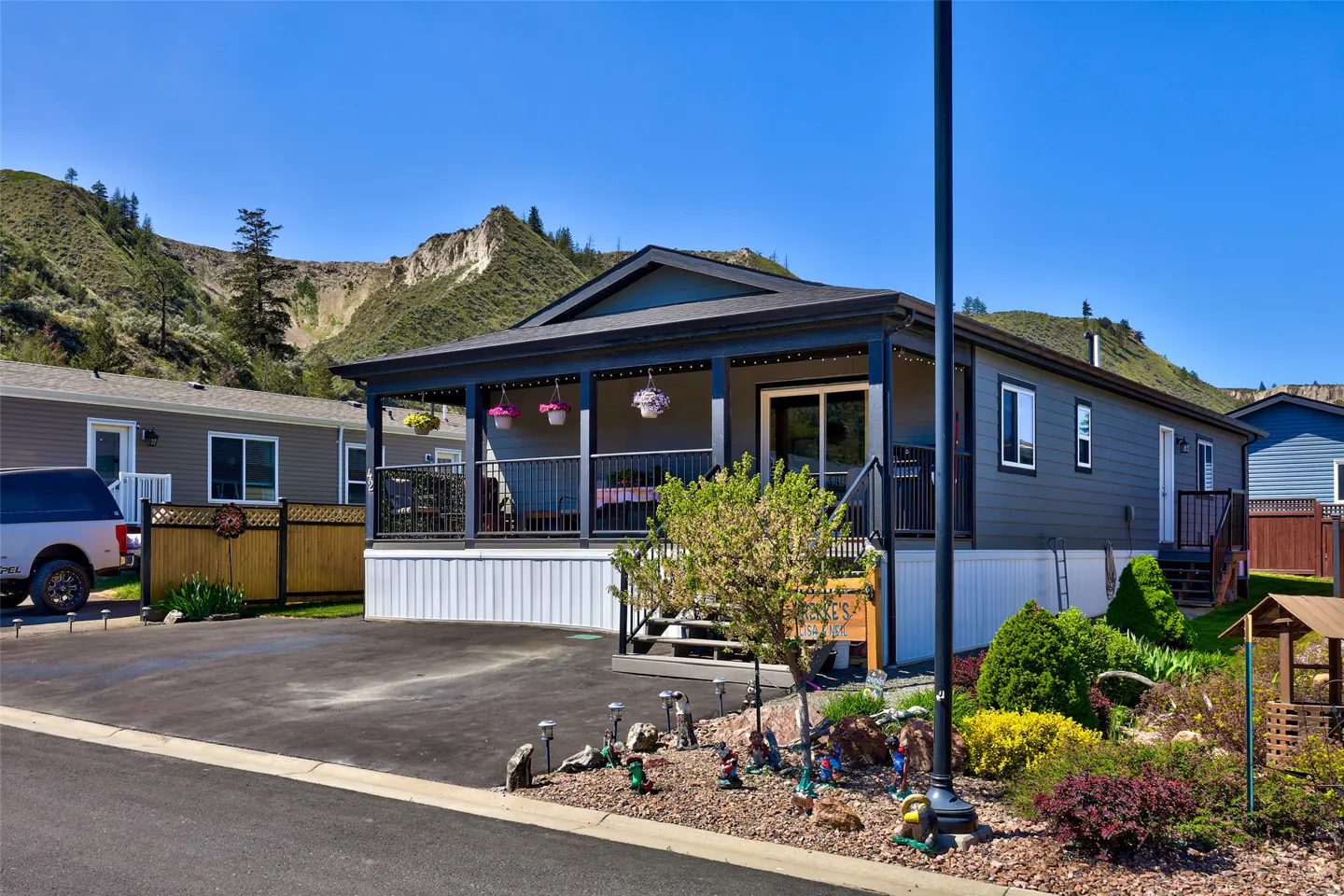 A gray mobile home with a covered porch and flower baskets, set against a mountain backdrop on a sunny day.