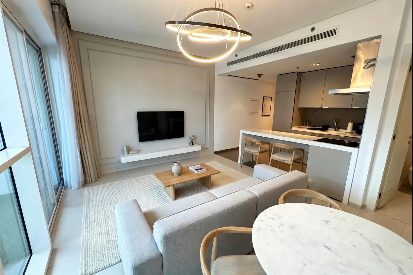 A modern apartment living space with a gray sofa, wood table, and open kitchen. A unique circular light fixture hangs above.