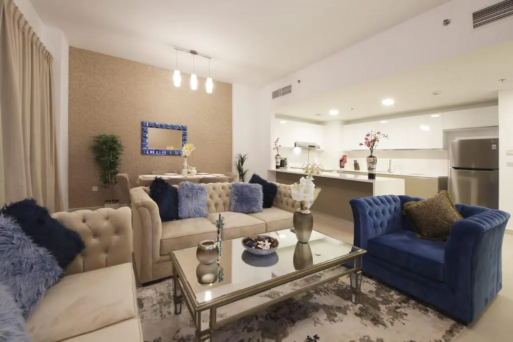 A modern living room with beige sofas, blue pillows, a glass coffee table, and an open kitchen in the background.