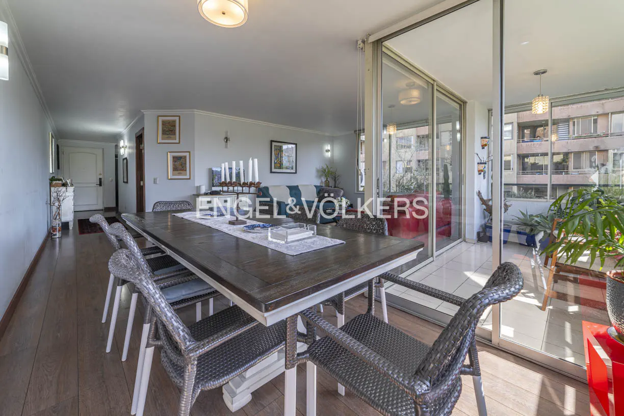 A dining room with a long wooden table and wicker chairs, leading to a balcony with city views.