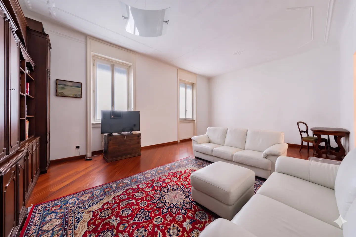 Living room with white leather sofas, a matching ottoman, a red patterned rug, and a dark wood cabinet on hardwood floors.