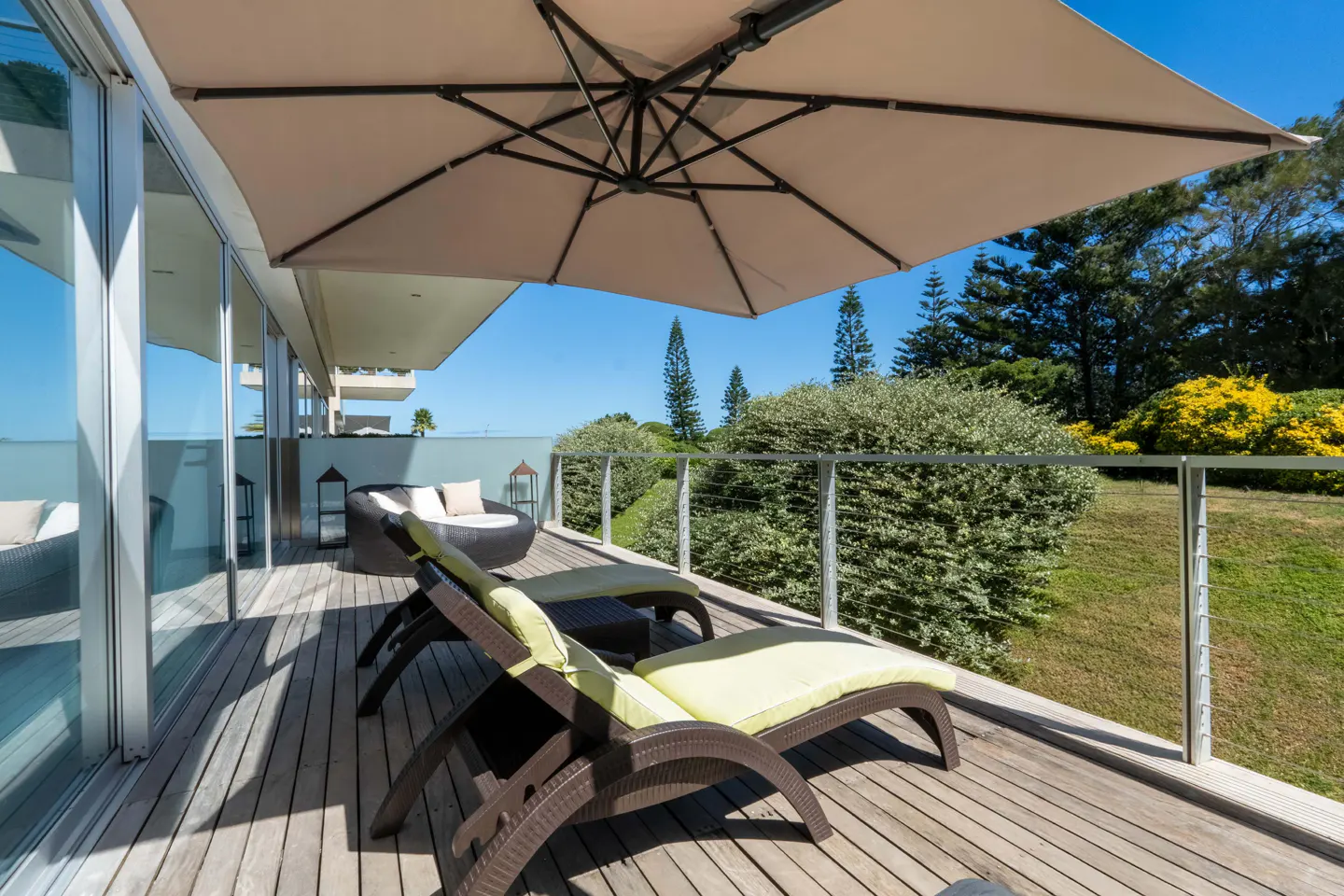 Wooden deck with lounge chairs and a beige umbrella. Green bushes and trees are in the background under a blue sky.