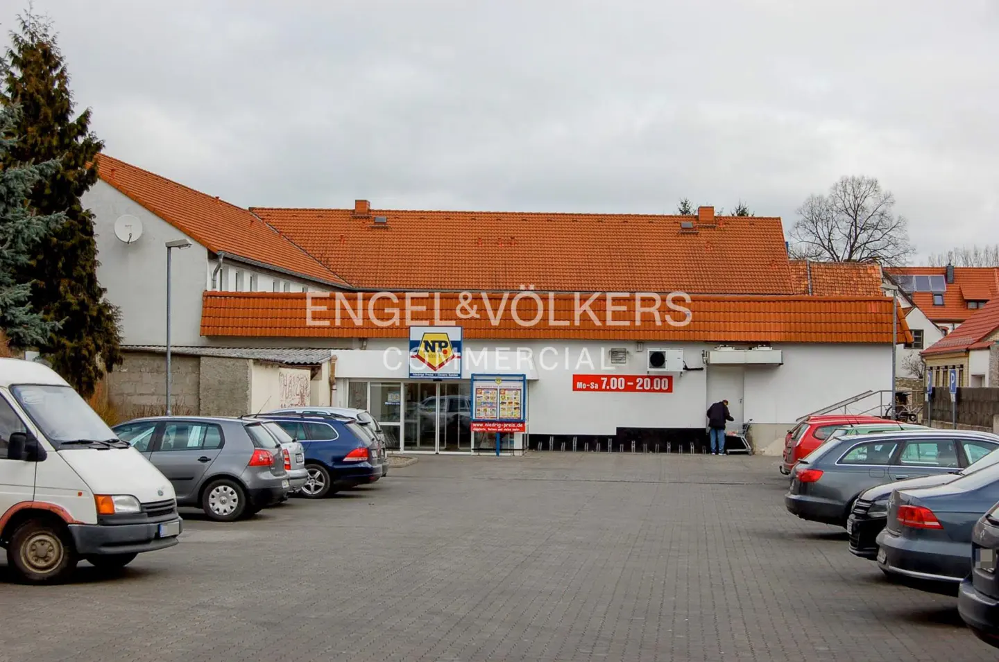 Exterior view of a white commercial building with an orange roof, Engel & Völkers signage, and a parking lot with cars.