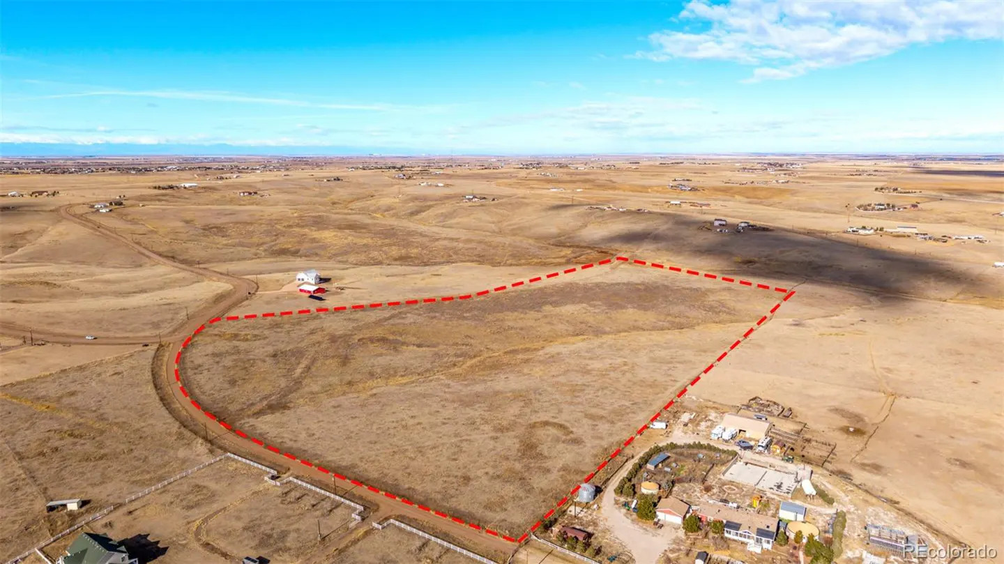 Aerial view of a large, vacant lot outlined in red, with sparse vegetation and scattered homes in the distance under a blue sky.