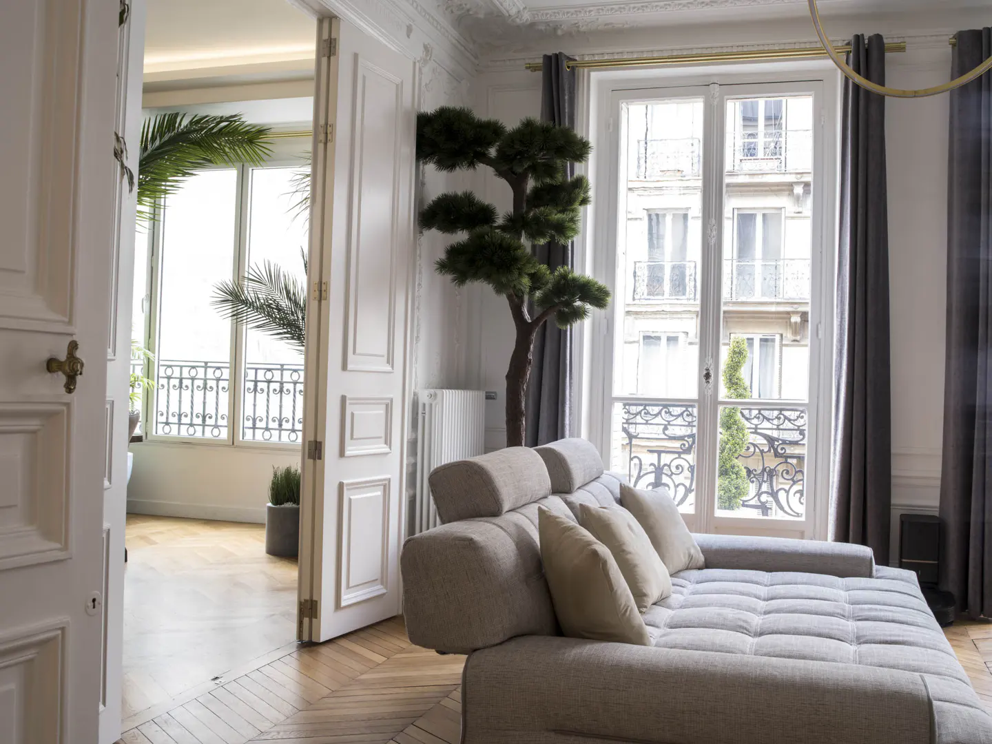 Bright living room with a beige sofa, a tall decorative tree, and double doors opening to a balcony with city views.