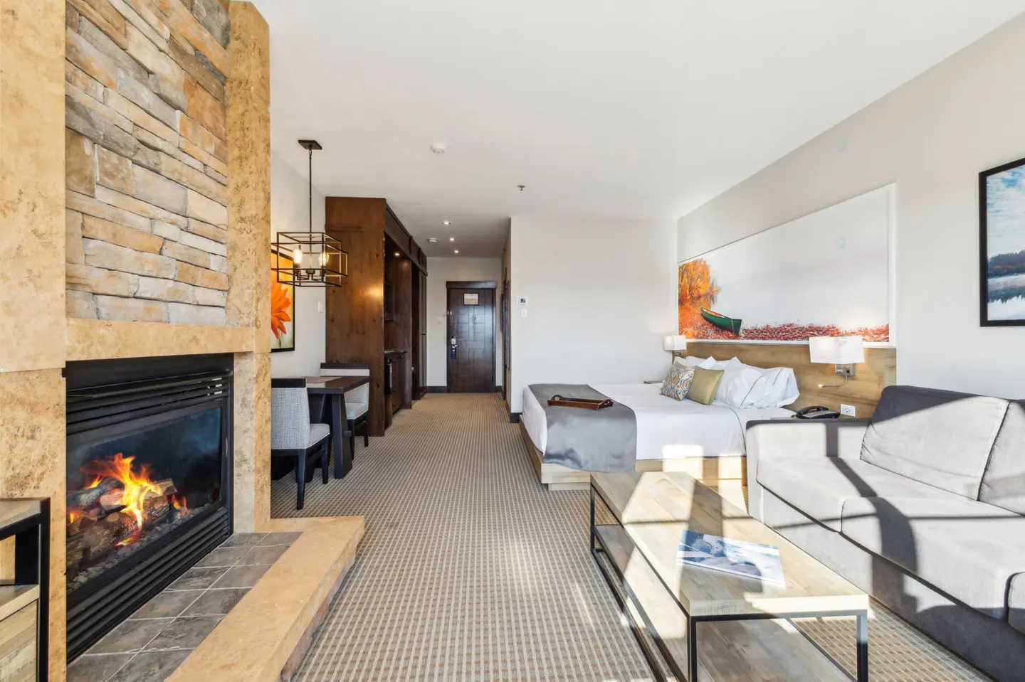 Hotel room with a stone fireplace, a bed with white linens, a gray sofa, and a wooden coffee table. A dining table and chairs are near a dark wood cabinet.