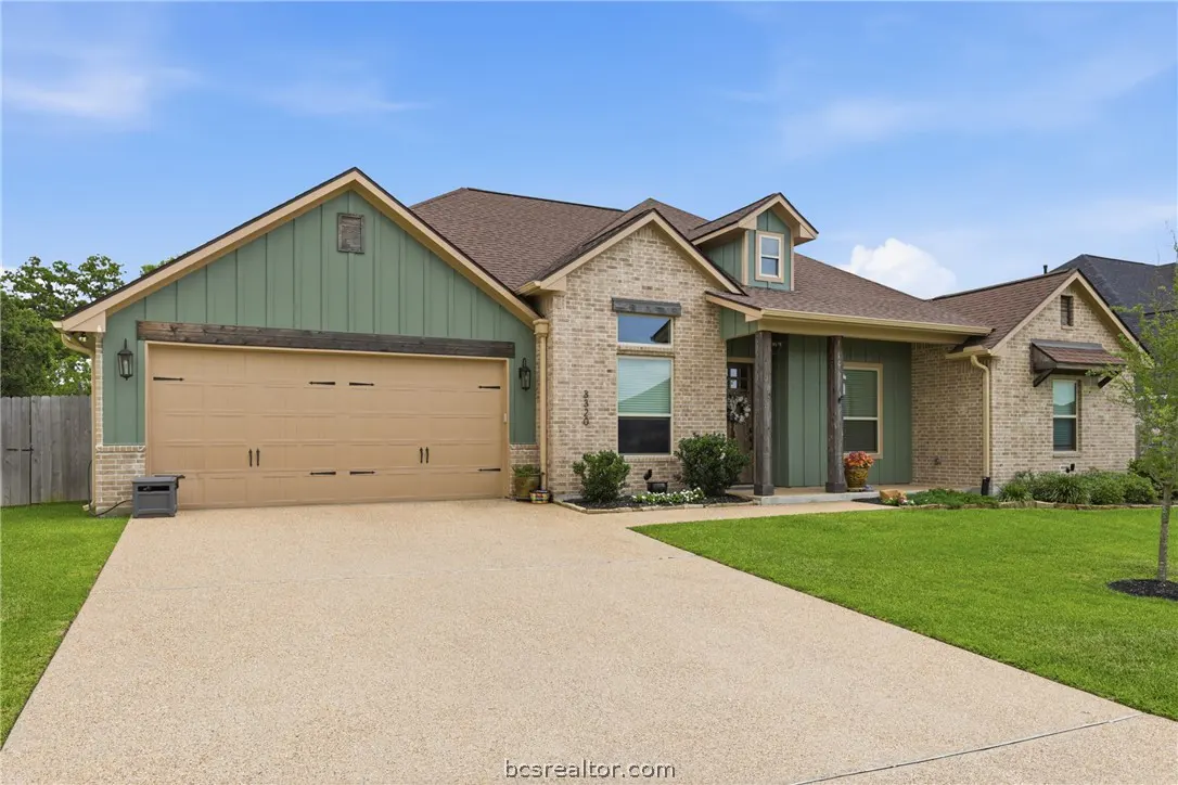 A single-story home with a tan brick and green siding exterior, a brown roof, and a beige driveway.