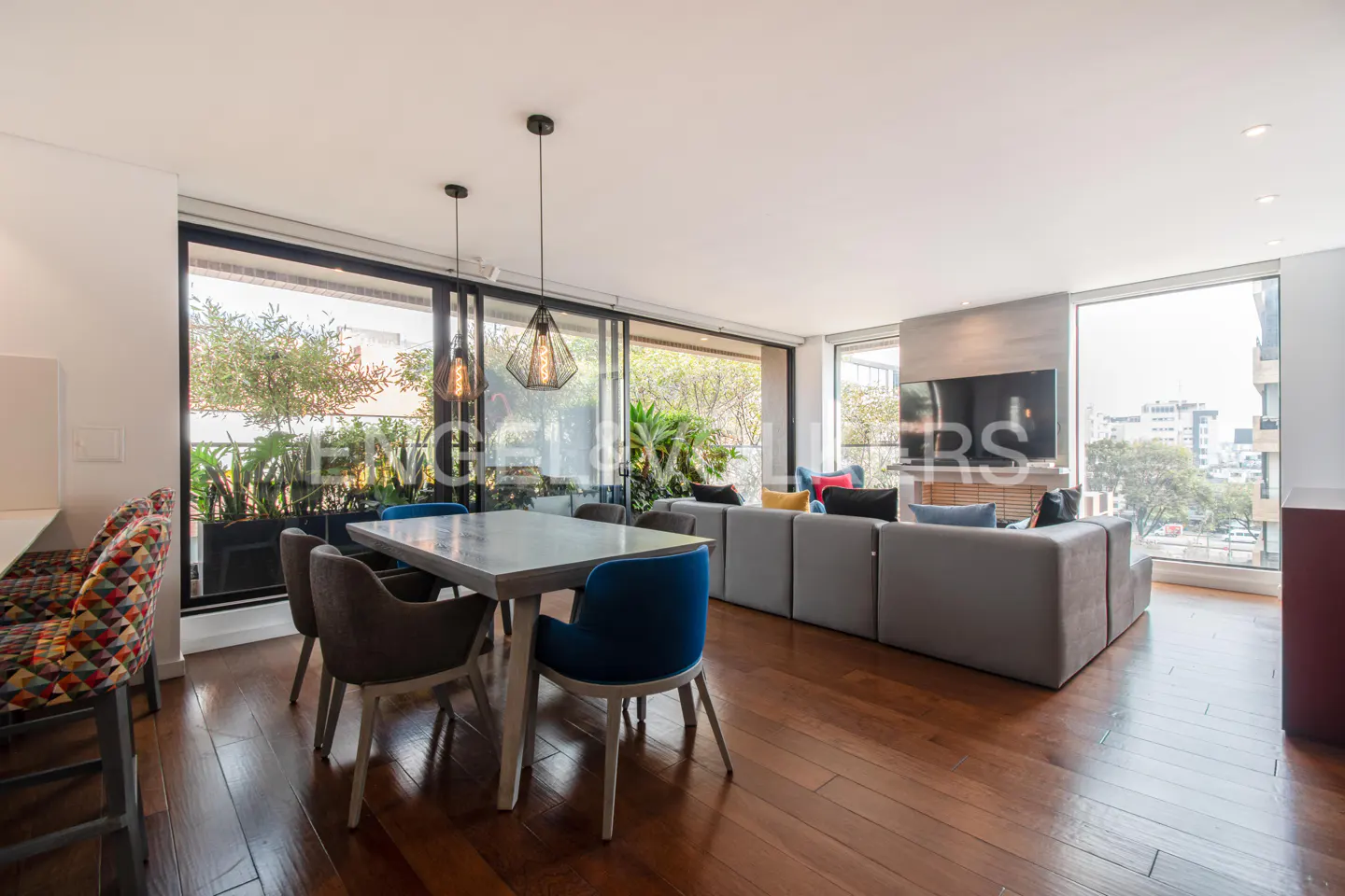Open-concept living space with wood floors, a dining table with blue and gray chairs, and a gray sectional sofa. Large windows offer city views.