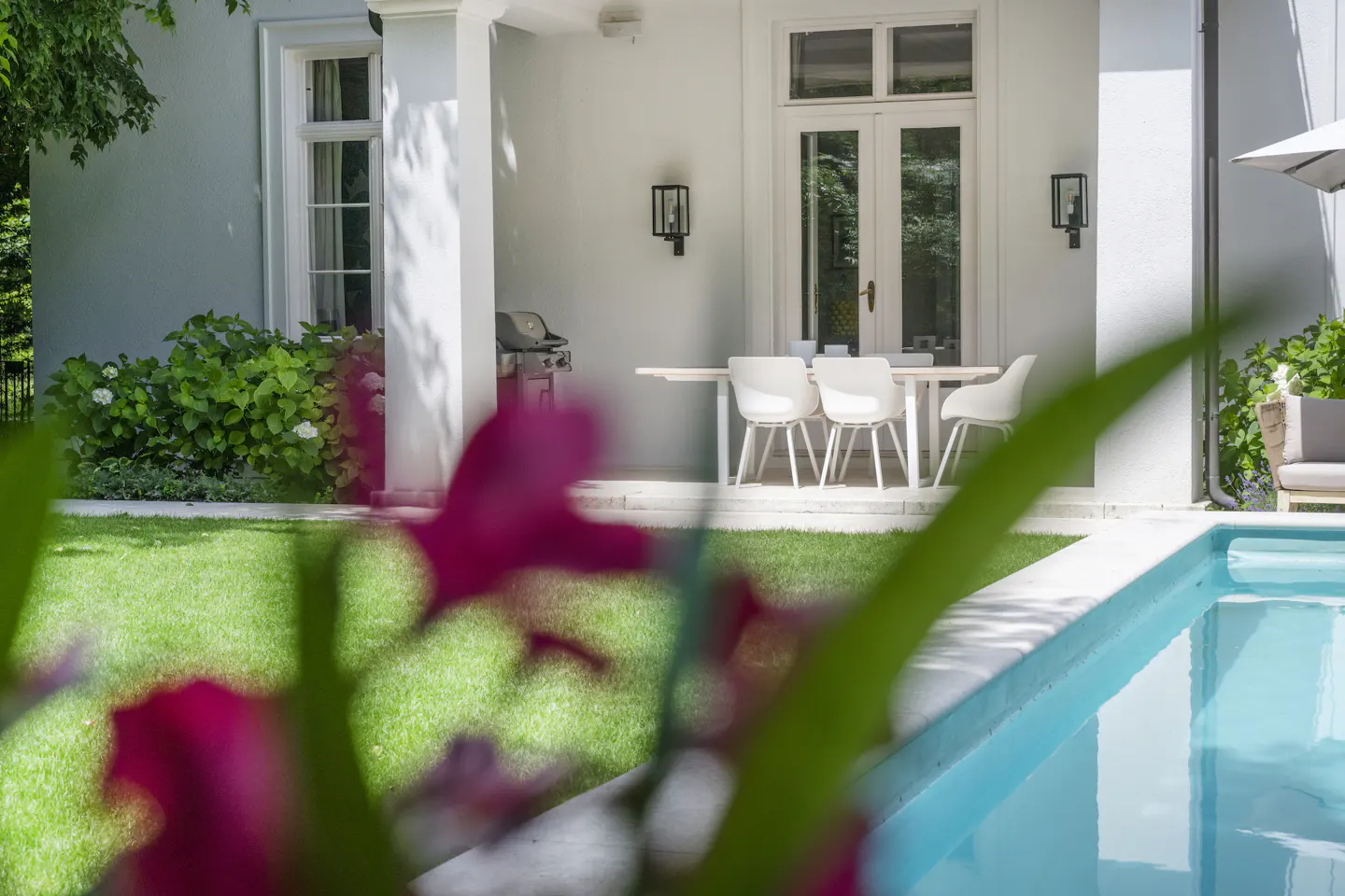 Outdoor patio with a pool, white table and chairs, grill, and green lawn. Pink flowers are in the foreground.