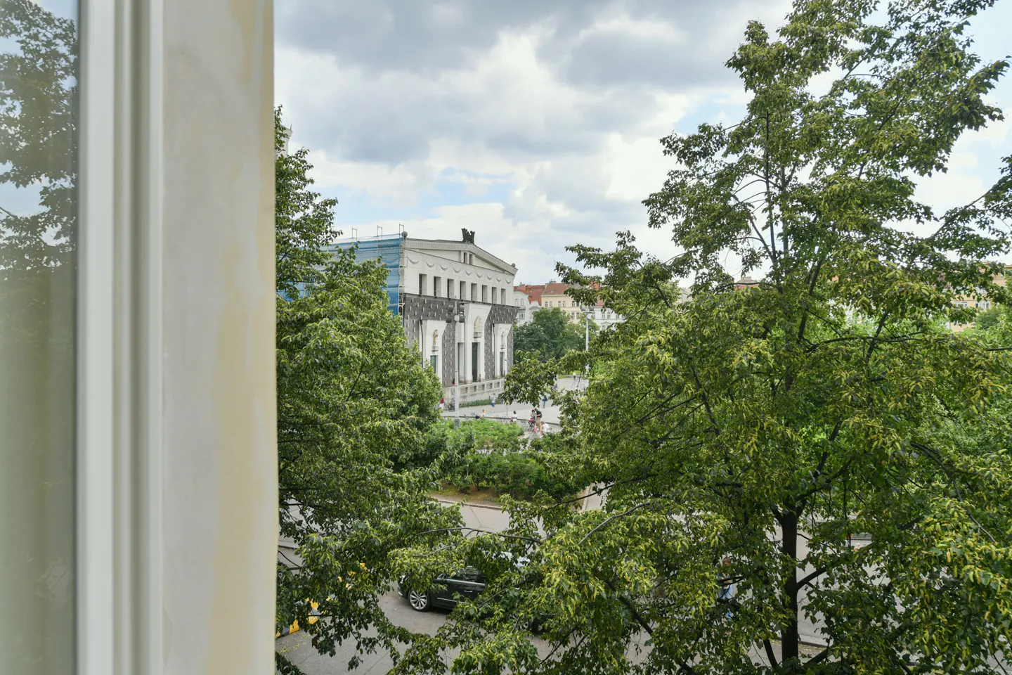 View from a window of a building across the street, partially obscured by green trees. Cloudy sky above.