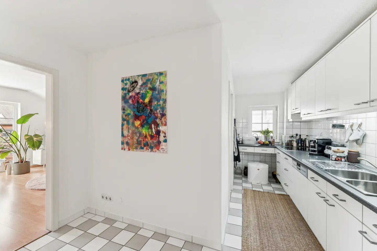 Bright, modern apartment hallway with a colorful abstract painting on a white wall, leading to a white kitchen.