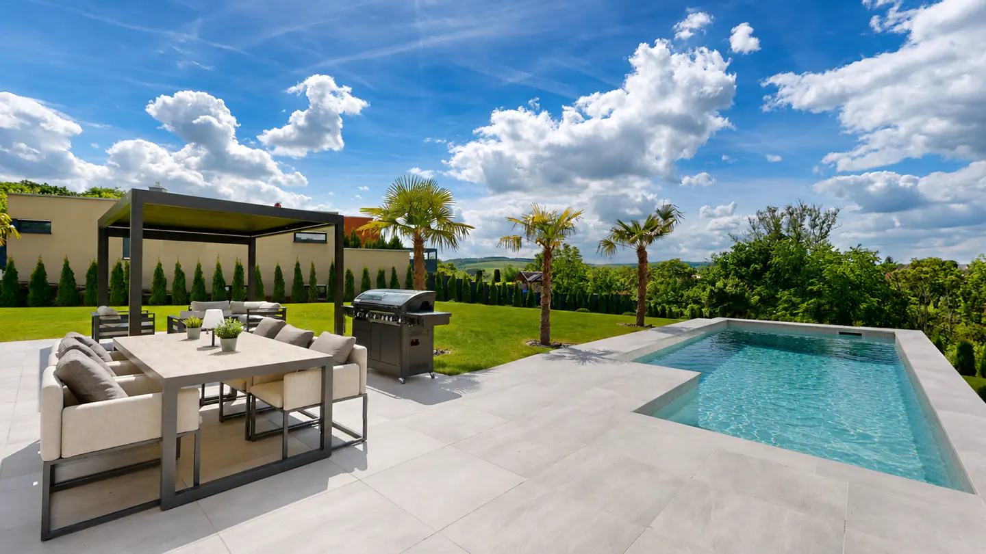 Outdoor patio with a pool, grill, and dining set. Palm trees and green lawn under a blue sky with white clouds.