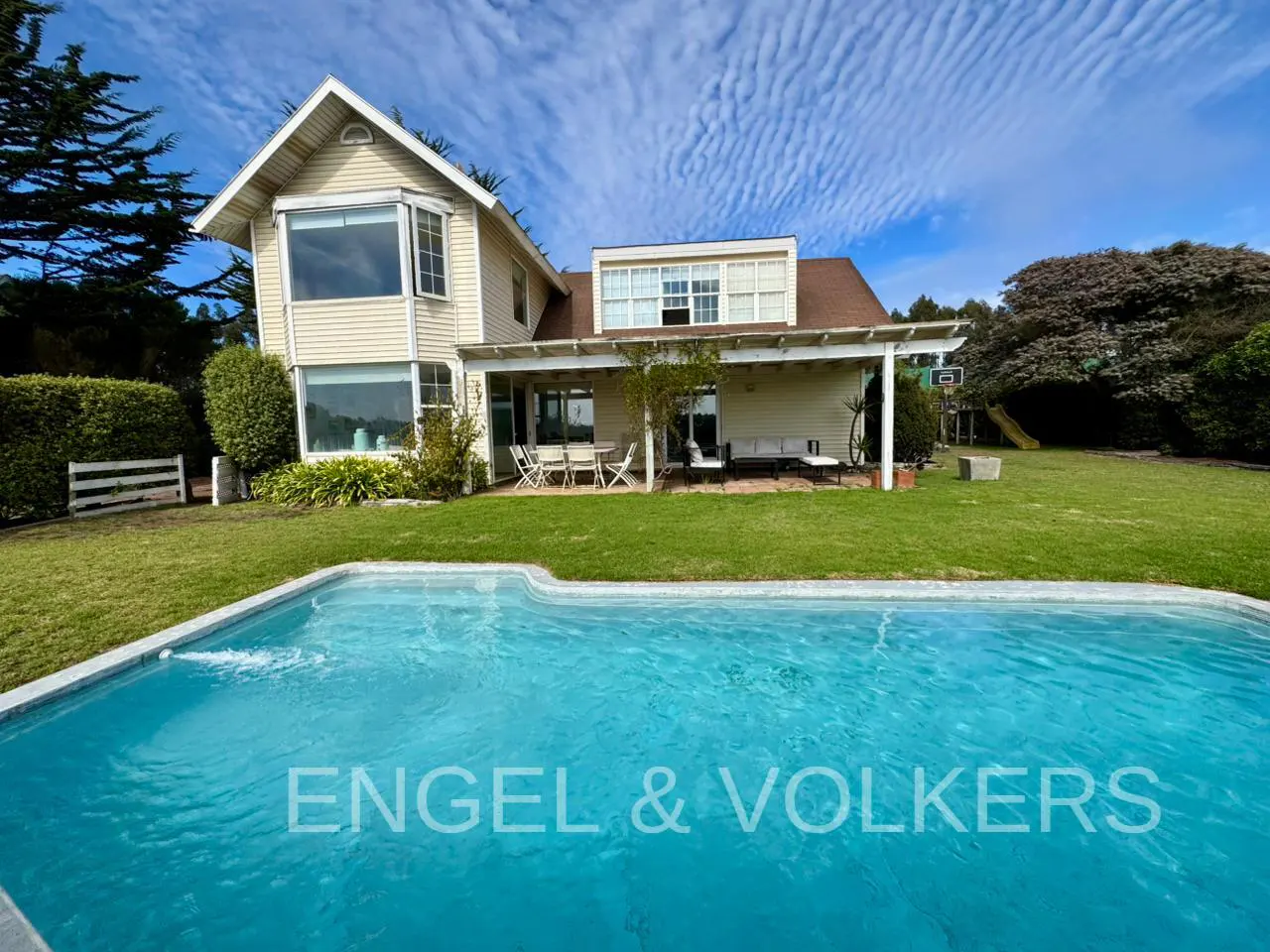 Exterior view of a two-story beige house with a pool, green lawn, patio furniture, and trees under a blue sky.