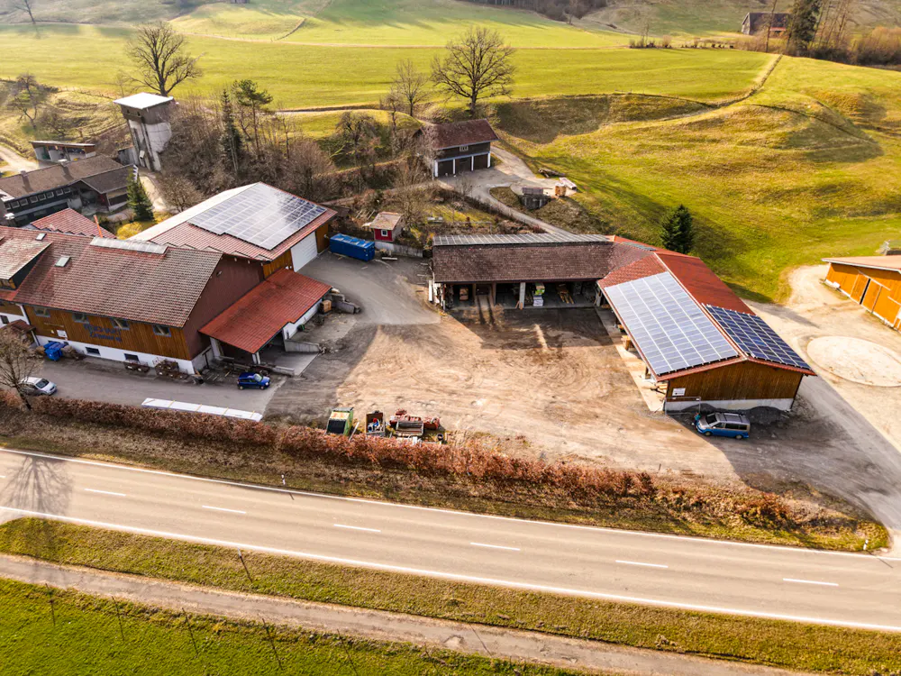 Aerial view of a farm with red-roofed buildings, solar panels, and a road in front, set against green fields and hills.