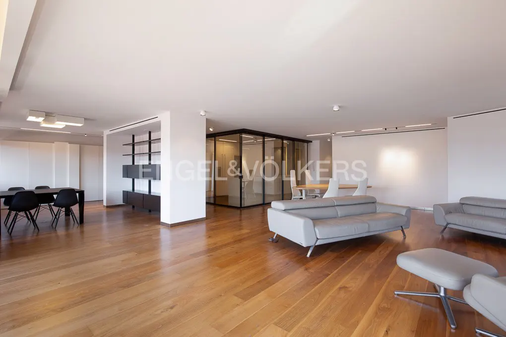 Bright, modern office space with wood floors, gray sofas, and a glass-walled conference room. A black table and chairs are on the left.
