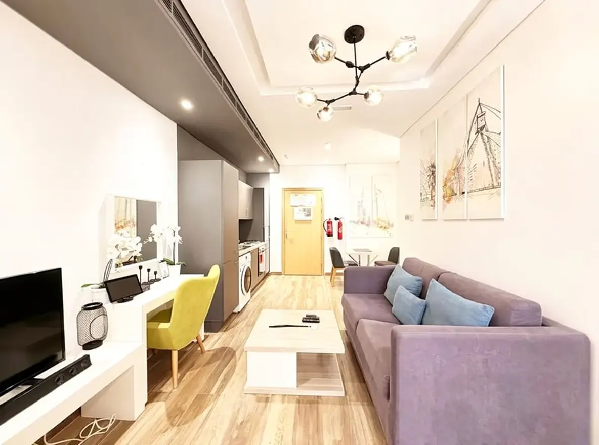 A modern apartment interior with a purple sofa, light wood floors, and a gray kitchen area. Artwork hangs on the white walls.