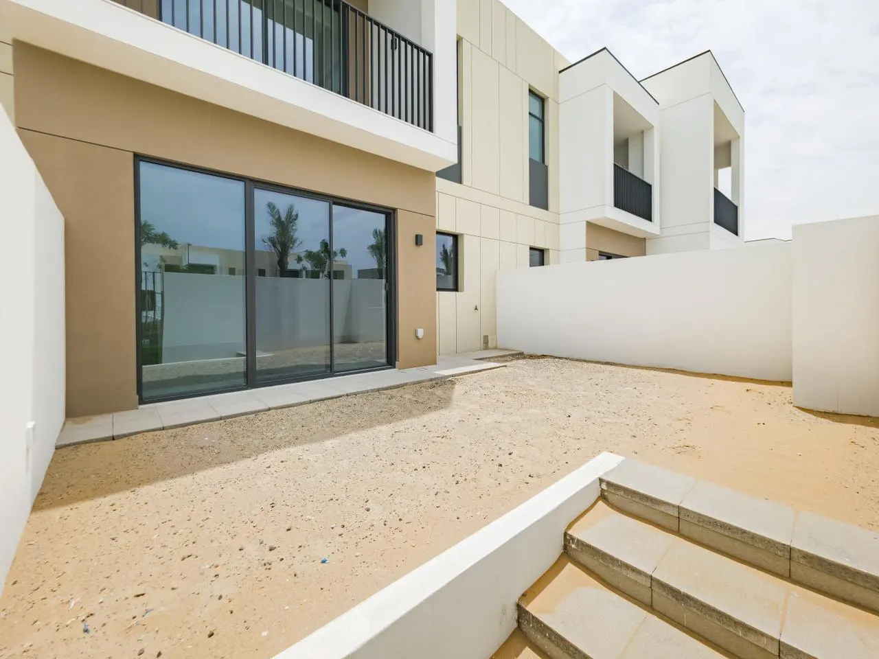 Exterior view of a modern two-story house with a sandy backyard and sliding glass doors. White walls surround the property.