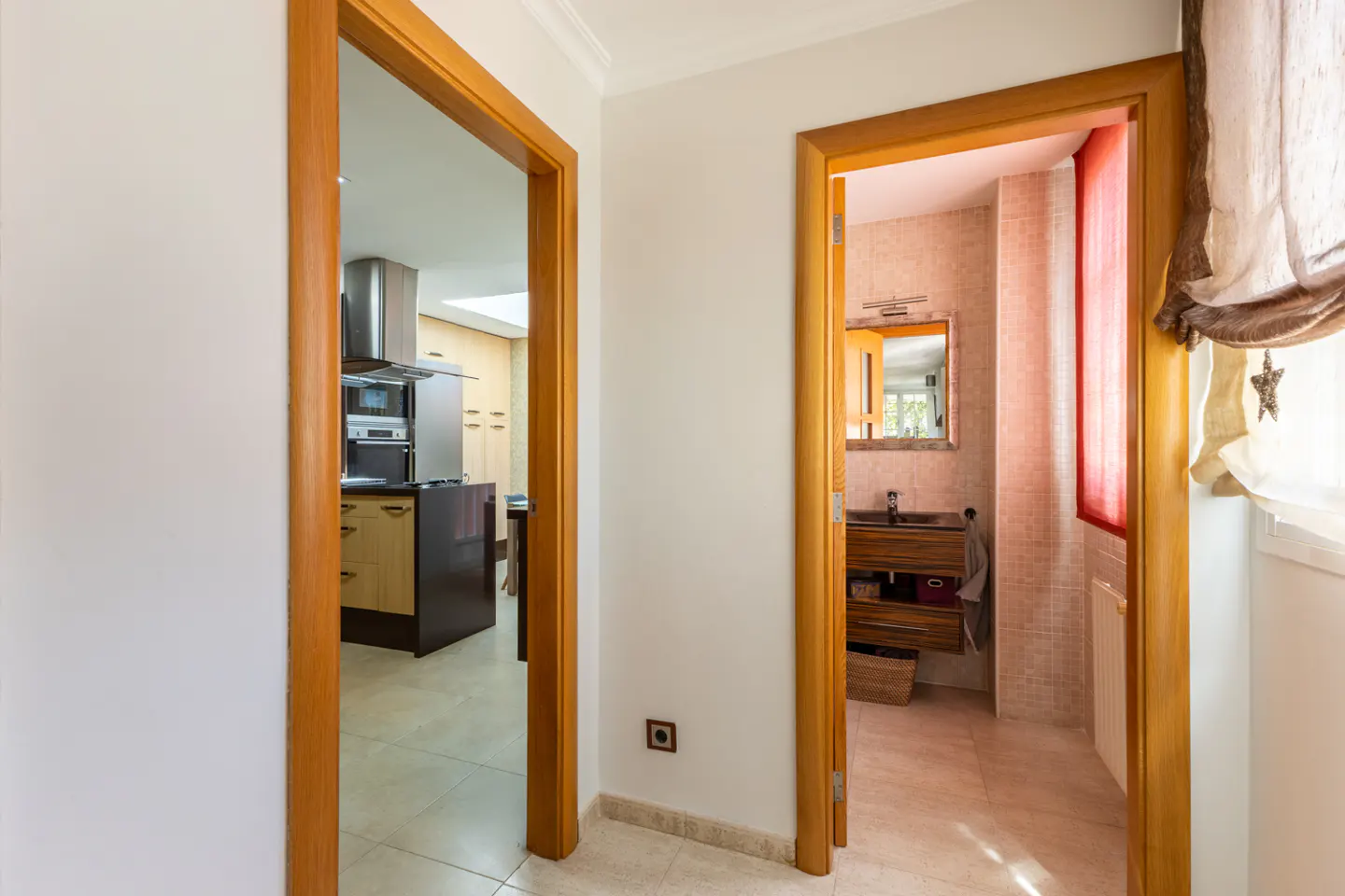 Hallway with two open wood-framed doorways. One leads to a kitchen, the other to a bathroom with a window and brown curtains.