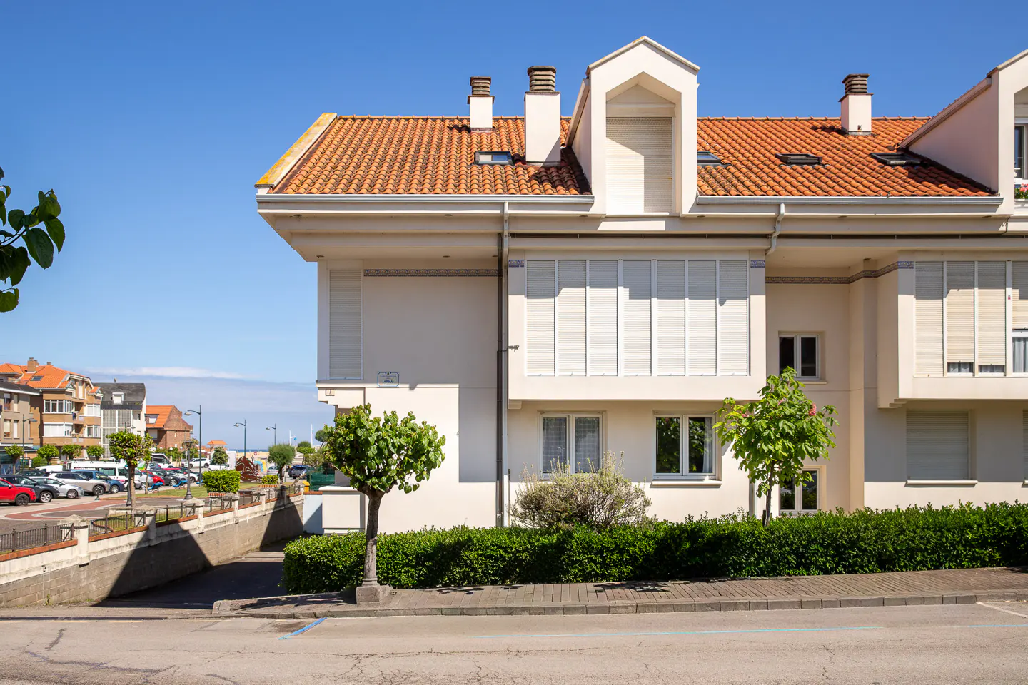 Exterior view of a modern, multi-story white building with a red tile roof, chimneys, and white blinds, set against a clear blue sky.