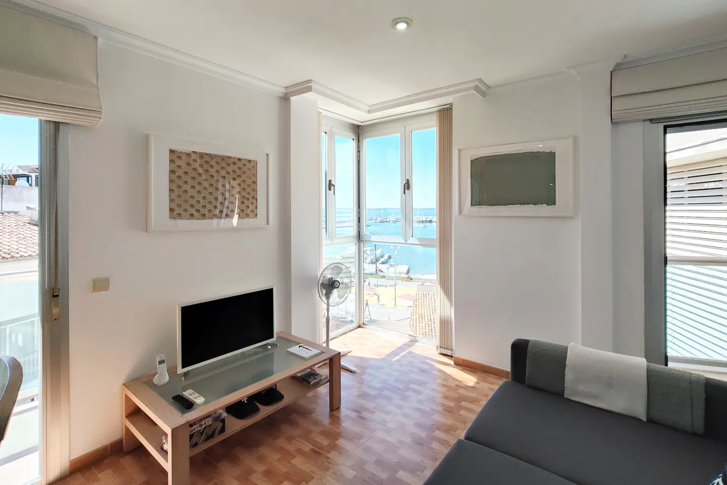 Bright living room with wood floors, white walls, and a view of the ocean through open windows. A gray sofa sits near a TV on a glass-topped table.