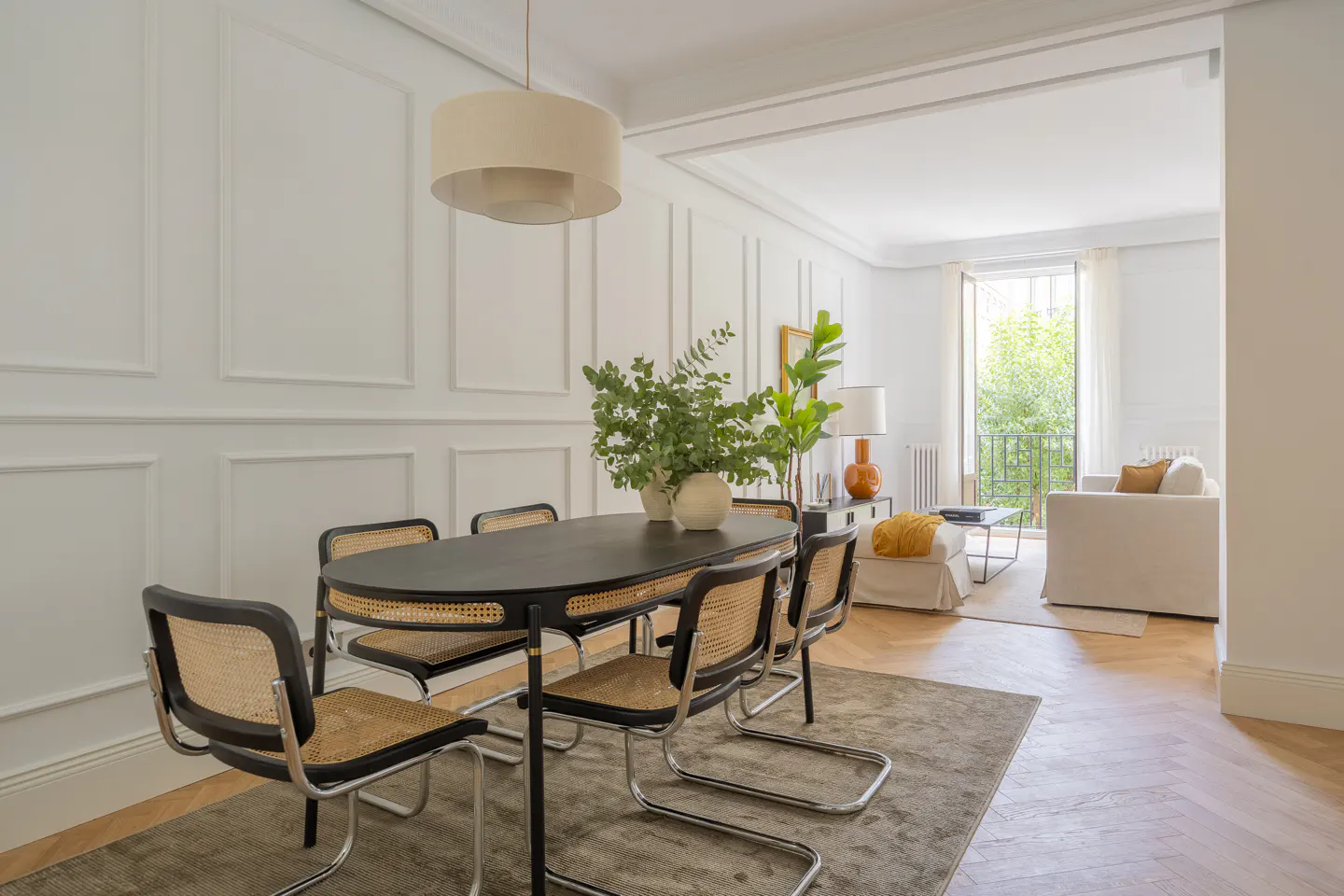 Modern dining room with oval black table, cane chairs, and white paneled walls, opening into a bright living area.